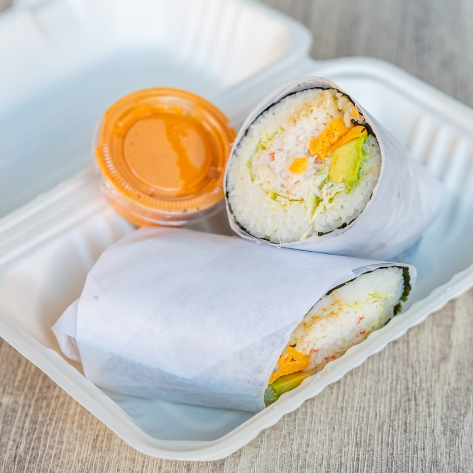 Two sushi burritos with crab, avocado, and orange sauce in a white takeout container.