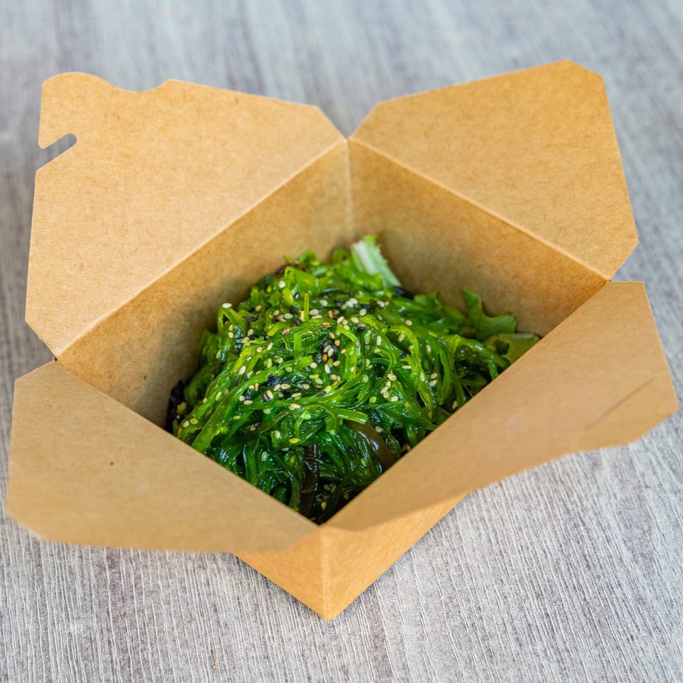 Ramen Kenjo's vibrant green seaweed salad with sesame seeds, served in a brown takeout container.