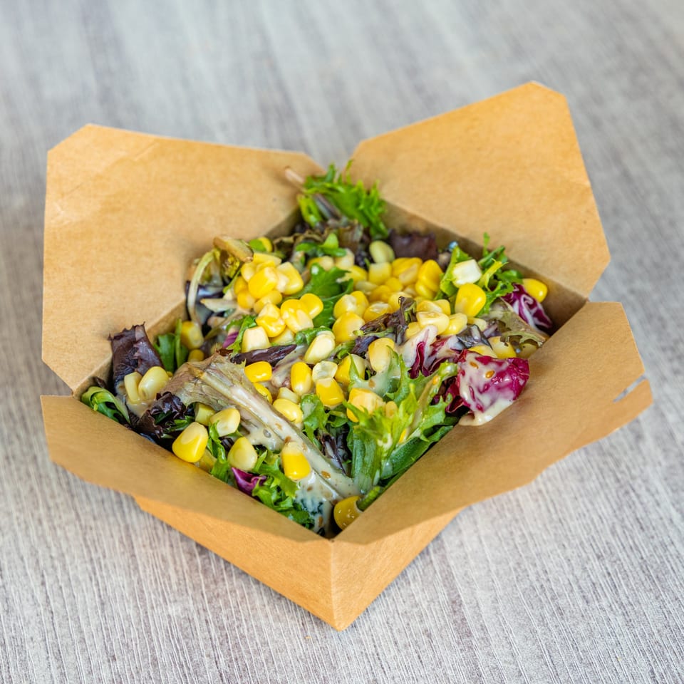 Fresh green salad with corn and dressing in a brown takeout container on a wooden table.