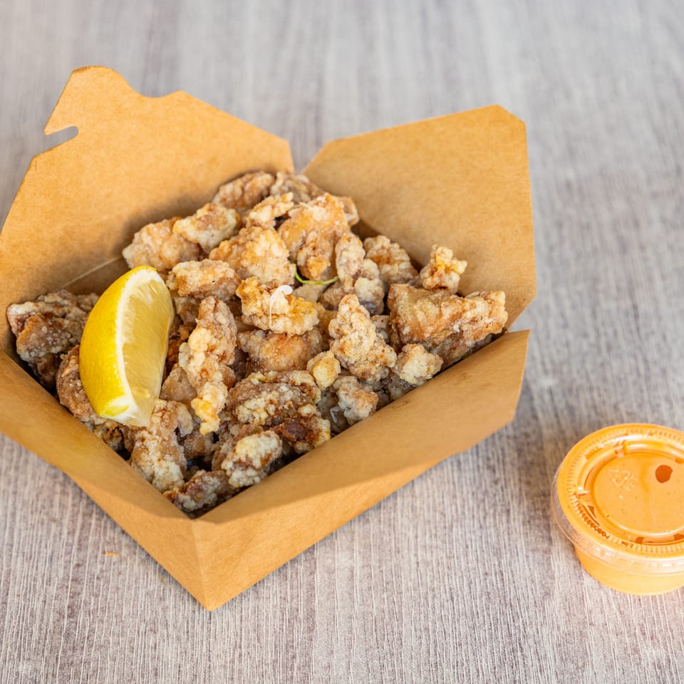 Crispy fried chicken karaage with a lemon wedge in a brown takeout box, with a side of sauce.
